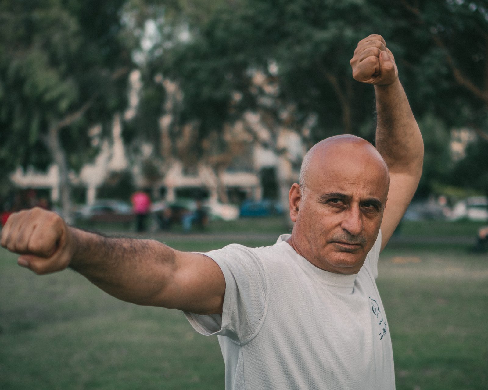 מתאמן בוגר בבית הספר וו מיי קונג פו מבצע צורת קונג פו בעמידה איתנה — אגרוף אחד פרוש לצד ואגרוף שני מורם מעל הראש, מבט ממוקד וקבוע. תרגול בפארק עם עצי איקליפטוס ונוף עירוני מטושטש ברקע.