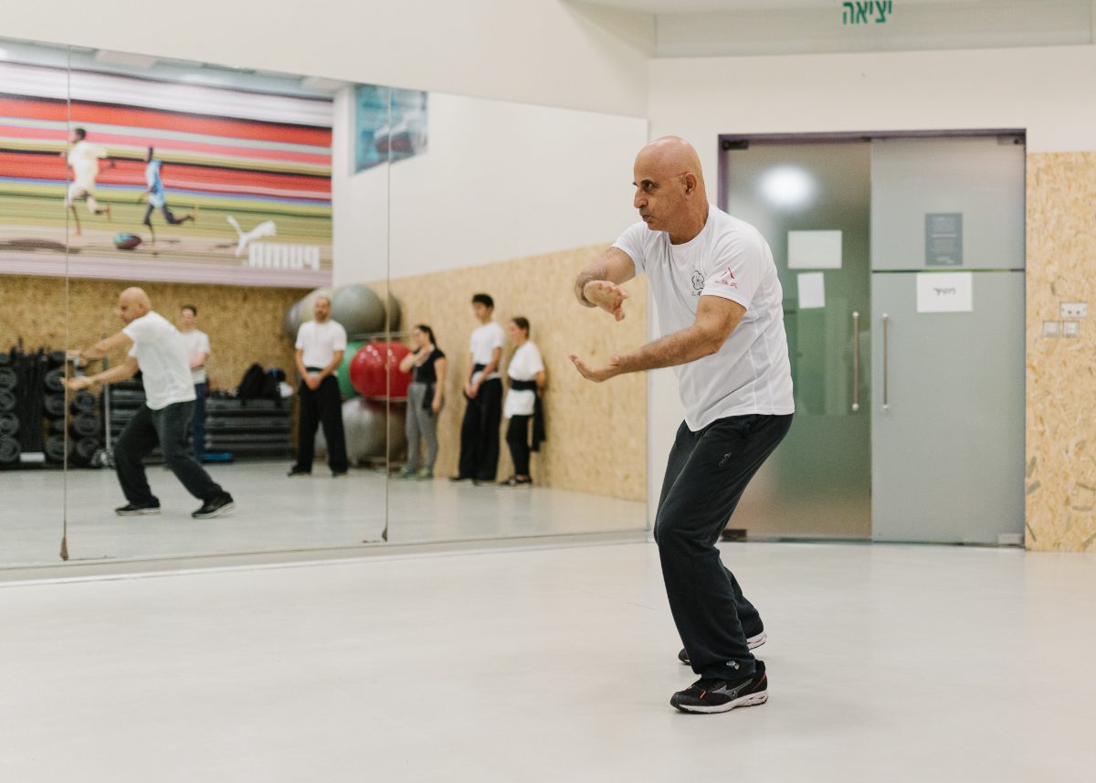 סיפו אבנר אקוע מבצע צורת קונג פו בסטודיו אלנבי 76 — עמידה נמוכה, ידיים מאוזנות לפנים בתנועה זורמת. ברקע מתאמנים צופים, ומראת הסטודיו משקפת את התנועה.