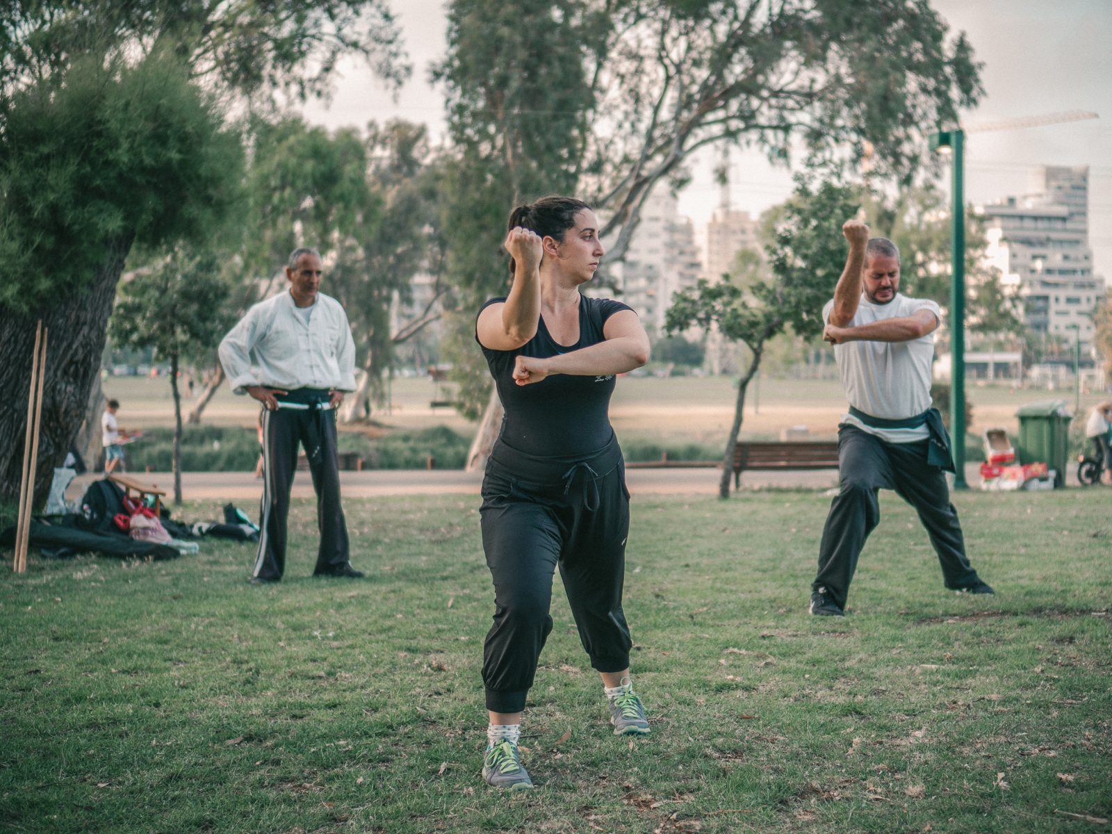 מתאמנת בולטת בקדמת התמונה מבצעת תנועת קונג פו בעמידה איתנה — אגרוף אסוף מול הפנים, מבט ממוקד. ברקע סיפו אבנר משגיח ומתאמן נוסף מבצע את אותה התנועה. אימון בפארק הירקון, עצי איקליפטוס ונוף עירוני ברקע.
