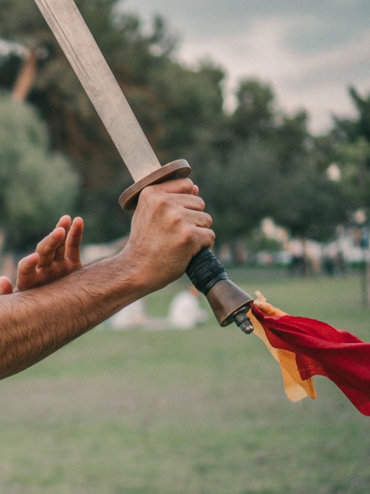 חרב רחבה (דאו / Broadsword) סינית מסורתית עם דגלון אדום־זהב — שתי ידיים אוחזות בחרב בפארק, סמל הוו מיי קונג פו